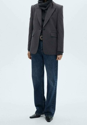 Personne portant un blazer gris foncé, un pull à col roulé noir, un jean bleu à jambes larges, une ceinture noire et des chaussures noires à bouts pointus, debout sur un fond clair.