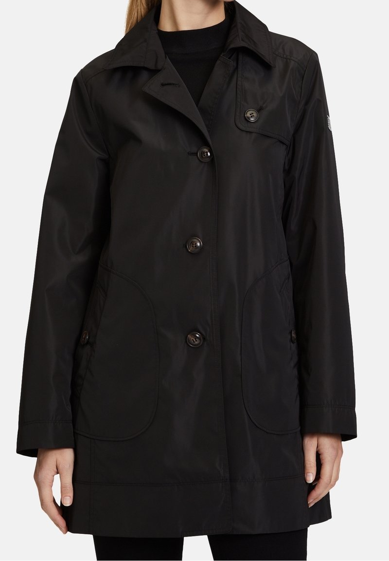 Femme portant un imperméable noir boutonné avec de grandes poches avant et un col, montrée du cou jusqu'au milieu de la cuisse sur un fond uni.