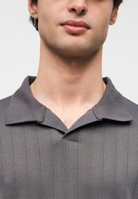 Polo gris avec une texture côtelée, un col plat et des lignes verticales. Le col présente une légère ouverture à l'avant.