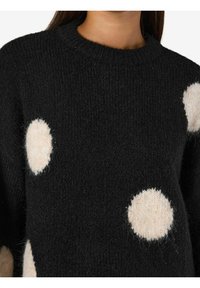 Suéter negro con lunares beige redondos, textura de punto, cuello acanalado y un corte general holgado. Material suave y cálido.