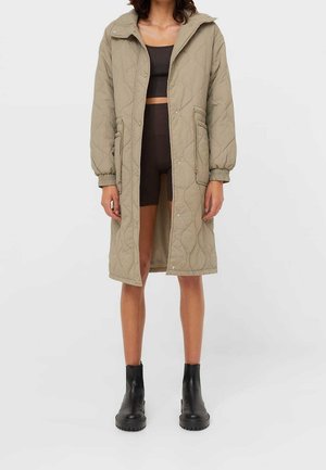 Vrouw die een lange beige doorgestikte jas draagt, een zwarte cropped top, zwarte biker shorts en zwarte enkellaarzen, staand tegen een effen achtergrond.