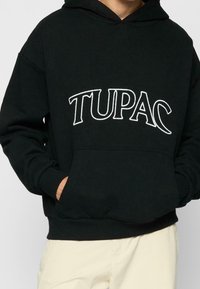 Svart hoodie i mjukt tyg med en framficka och vit text "TUPAC" tvärs över bröstet. Standardpassform med ribbade ärmslut.