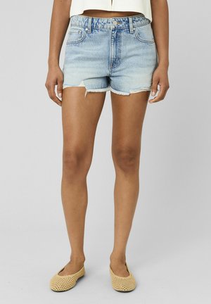 Femme portant un short en jean bleu clair coupé avec des bords effilochés et des chaussures plates tressées beige, debout devant un fond neutre.