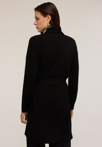 Cardigan noir à manches longues avec une taille ceinturée, présentant des accents texturés aux épaules. Tissu doux et lisse avec un design épuré.