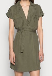 Abito verde khaki con maniche corte, scollatura a V profonda, tasche frontali e dettaglio della cintura. Realizzato in tessuto leggero e liscio.