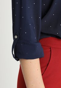 Manche de chemise bleu marine à petits pois blancs roulée au-dessus du coude, associée à un pantalon rouge taille haute avec poche latérale.