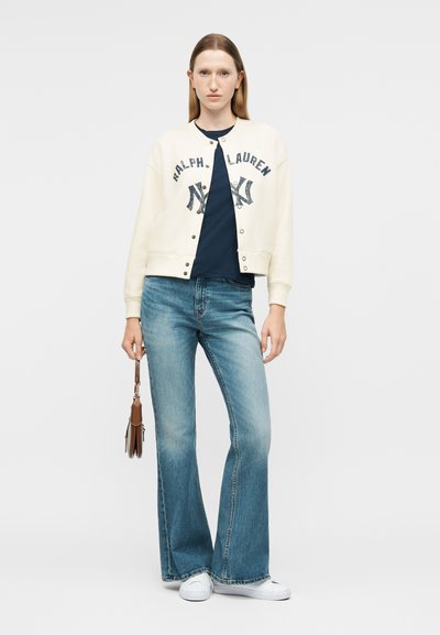 Γυναίκα που φοράει κρεμ ζακέτα Ralph Lauren, μπλε σκούρα μπλούζα, καμπάνα μπλε τζιν, λευκά αθλητικά παπούτσια, κρατώντας μια μικρή καφέ τσάντα στο πλάι της.