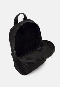 Černý látkový batoh se zipovým uzávěrem, prostorným vnitřkem a jedním držadlem na nošení. Obsahuje polstrované části pro lepší organizaci.