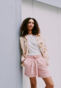 Lys beige jakke over en hvit ribbet topp, sammen med rosa shorts med snøring i livet. Bakgrunnen er enkel og nøytral.