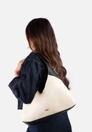 Femme aux longs cheveux bruns portant une veste en jean foncée et un grand sac à bandoulière en cuir blanc et noir.