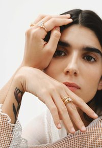 Anillos de oro con diseños intrincados, incluyendo una gran gema rectangular, llevados en una mano con un tatuaje floral. Manga de malla visible.