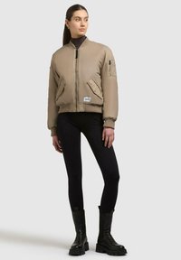 Beige Bomberjacke mit Reißverschlussfront, gerippten Bündchen, zwei Fronttaschen und einer Seitentasche. Kombiniert mit schwarzen Leggings und Stiefeletten.