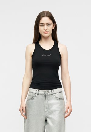 Mujer con cabello largo y castaño, vestida con una camiseta negra sin mangas con el texto "diesel" y unos vaqueros plateados de talle alto, de pie frente a un fondo sencillo.