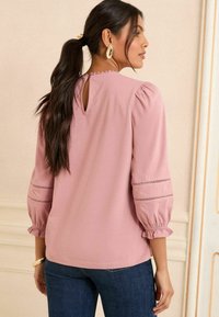 Rosa Bluse mit puffartigen Ärmeln, die über einen Schlüsselloch-Ausschnitt am Rücken verfügt und bestickte Details an den Ärmeln aufweist. Weicher Stoff mit glatter Textur.