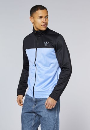 Mann steht, trägt eine schwarz-hellblaue Trainingsjacke mit Reißverschluss und Logo auf der Brust sowie blaue Jeans, schaut nach links.