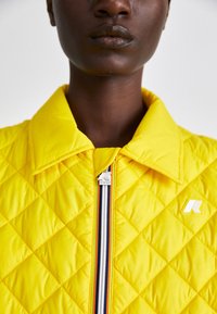 Casaco acolchoado amarelo com colarinho pontudo, fecho de zip na frente e um logótipo. Apresenta detalhes em listas perto do fecho e uma textura suave.