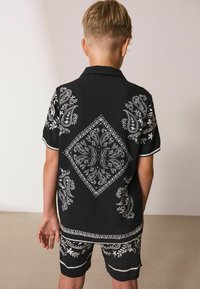 Čierna polokošeľa s krátkym rukávom a šortky s zložitými bielymi paisley a kvetinovými vzormi, s diamantom tvarovaným dizajnom na chrbte.