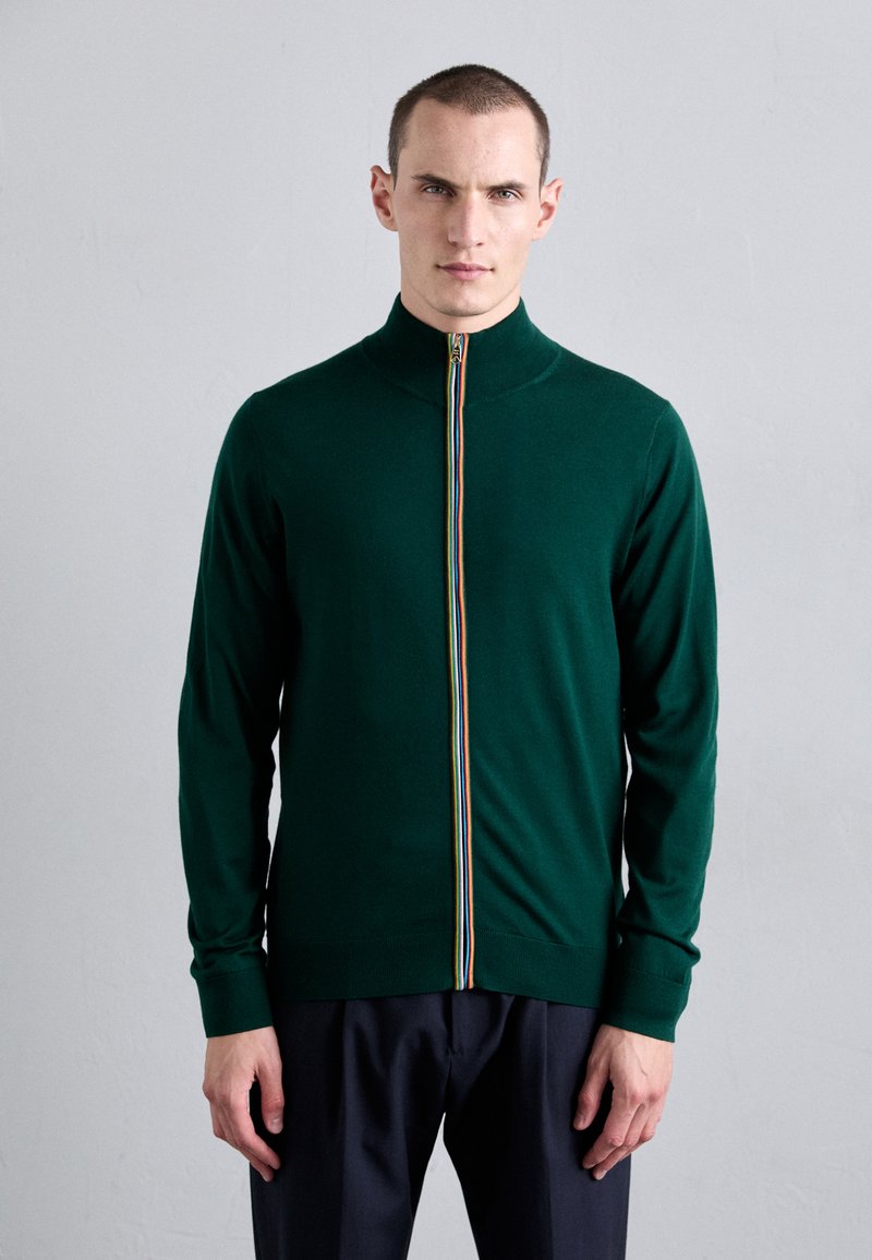 Grønn zip-up genser med stående krage, med en flerfarget stripe langs glidelåsen, laget av et glatt, elastisk strikket stoff.