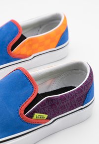 Vans slip-on skor i blå mocka med orange rutmönster, lila detaljer och elastiska sidogömmor. De har en vit yttersula och branding.
