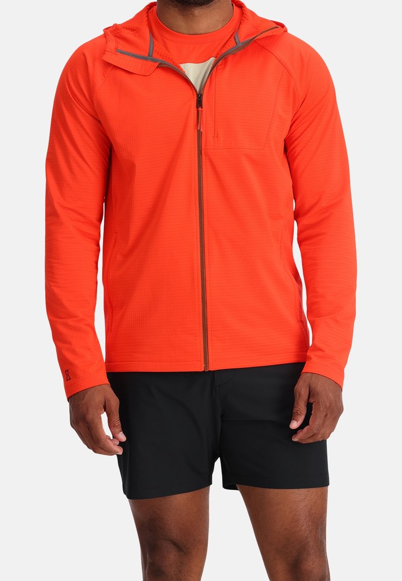 Homme portant une veste zippée orange vif et un short noir, debout devant un fond blanc uni.