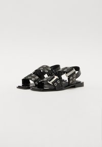 Black leather sandals featuring multiple adjustable straps with silver hardware and circular cutouts. Flat sole with a smooth texture.