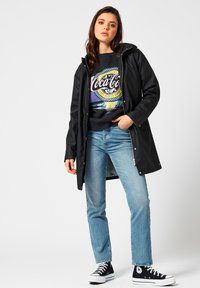 Imperméable noir et waterproof avec capuche, porté sur un sweatshirt graphique arborant des éléments colorés de la marque Coca-Cola, associé à un jean bleu clair et des sneakers noires.