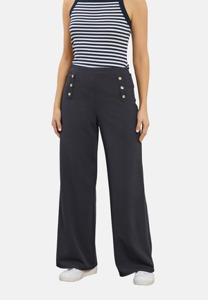 Kvinde iført marineblå bukser med vide ben og guldknapper på lommerne, kombineret med en ærmeløs top i marineblåt og hvidt stribet mønster samt hvide sneakers.