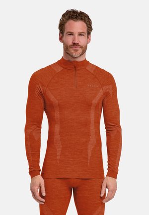 Mann steht, trägt eng anliegendes rustfarbenes thermisches Langarmshirt und passende Hose mit dezenten Musterdetails und Halb-Reißverschluss am Kragen.