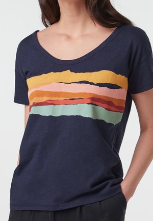 Mujer con una camiseta azul marino con un patrón de franjas horizontales multicolores en amarillo, rosa, naranja, rojo y verde claro a lo largo del pecho.