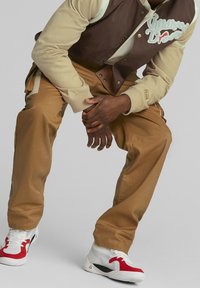 Brown and beige varsity jacket with embroidered text, paired with tan pants and white sneakers accented with red. Smooth fabric texture.