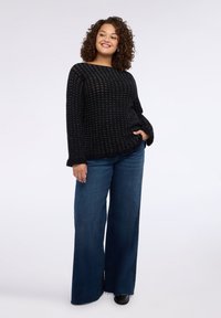 Černý pletený svetr s otevřeným designem, kombinovaný s širokými džínami a černou obuví. Přírodní kudrnaté vlasy rámují obličej.