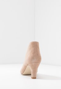 Beige suède hak schoen met een taps toelopend ontwerp, voorzien van een lage blokhak en een gladde textuur, geplaatst op een lichte achtergrond.