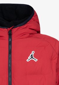 Czerwona kurtka puchowa z kapturem, czarną podszewką, zamkiem z przodu oraz białym logo Jumpman na piersi. Gładki, izolowany materiał.
