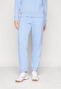 Pantalons de survêtement bleu clair en tissu doux et lisse avec des poignets élastiques aux chevilles et une coupe décontractée, associés à des chaussures de sport blanches.