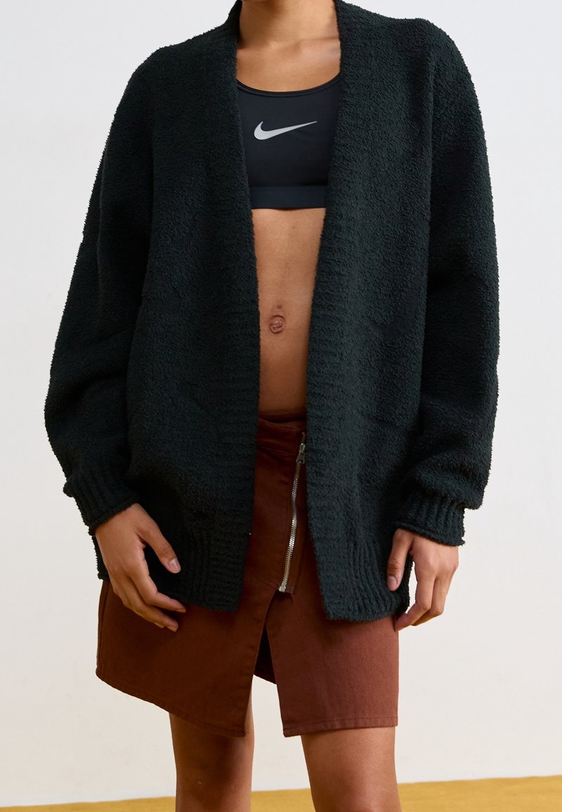 Person som bär en svart fluffig cardigan över en svart Nike sport-bh och en brun kjol med en dragkedjesprund framtill, stående mot en slät vägg.