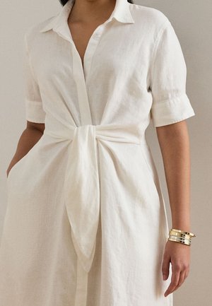 Kvinde iført hvid linnedkjole med korte ærmer og et bindedetalje foran, accessoriseret med tre gyldne armbånd på højre håndled.