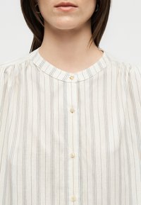 Gros plan d'une femme portant un chemisier blanc à fines rayures verticales bleu et orange, avec un col rond et des boutons sur le devant.