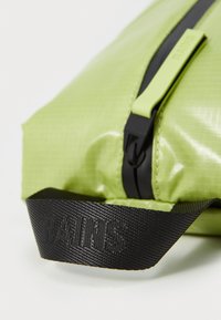 Saco impermeável verde com um acabamento texturizado, apresentando um fecho preto e uma alça preta com marcação em relevo. Inclui elementos de design ergonômico.