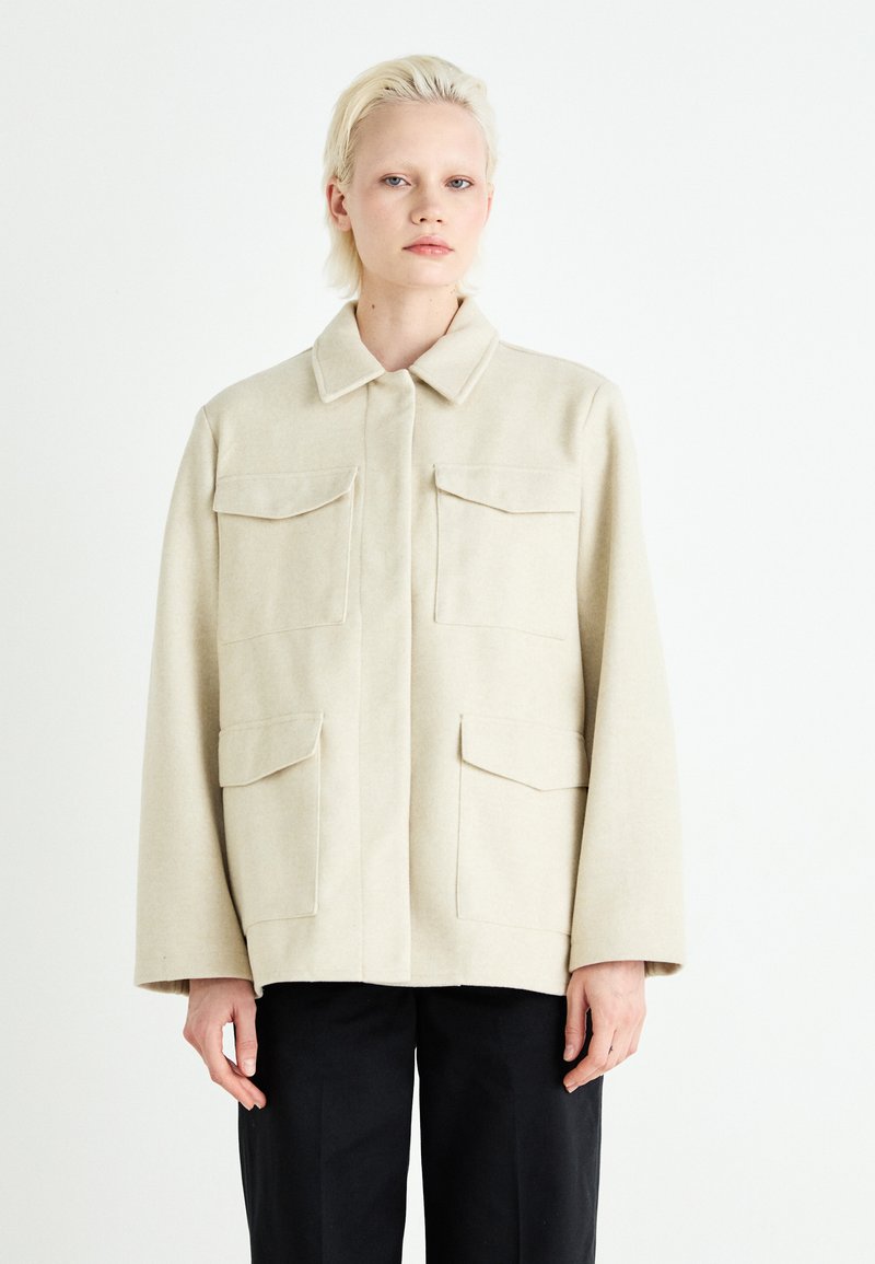 Beige ullblandingsjakke med fire frontlommer, krage og lange ermer. Stoffet har en myk tekstur og en strukturert design.