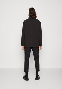 Maglietta nera a maniche lunghe con taglio dritto, abbinata a pantaloni scuri e scarpe nere, vista da dietro su uno sfondo bianco semplice.