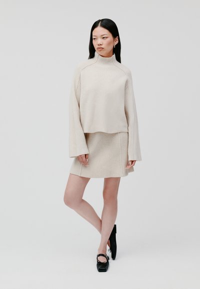 Pullover a maglia rigata beige con collo alto e maniche ampia, abbinato a una gonna mini rigata coordinata. Indossato con scarpe nere.
