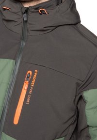 Šedá a zelená izolovaná bunda s kapsou na zip na hrudi s oranžovými detaily a nastavitelnou kapucí. Texturovaná látka, moderní design.