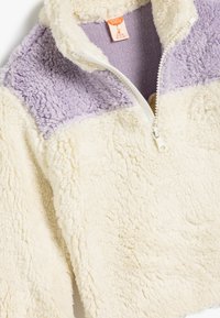 Pull en polaire crème et lilas avec un zip half, présentant une surface texturée et des blocs de couleur contrastants sur les épaules.
