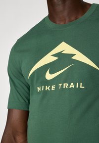 T-shirt en coton vert avec un grand motif de montagne jaune et le logo Nike, le texte "NIKE TRAIL" en dessous. Manches courtes, coupe décontractée.