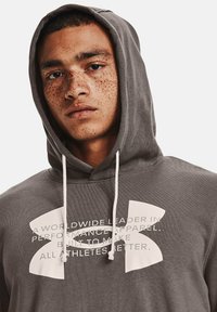 Grijze hoodie met een wit logo en tekst, voorzien van trekkoorden en een ontspannen pasvorm. De stof lijkt zacht met een gladde textuur.