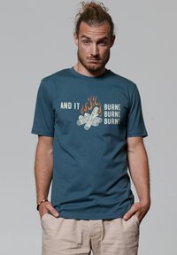Teal katoenen t-shirt met korte mouwen, voorzien van een graphic van brandende takken en vlammen, met de tekst "EN HET BRANDT, BRANDT, BRANDT."