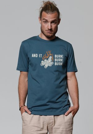 Teal katoenen t-shirt met korte mouwen, voorzien van een graphic van brandende takken en vlammen, met de tekst "EN HET BRANDT, BRANDT, BRANDT."