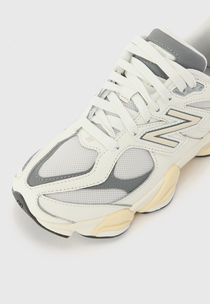 Scarpa sportiva bianca e grigia con sovrapposizioni in rete e materiale sintetico, dettagli testurizzati, colletto imbottito e logo distintivo sul lato.