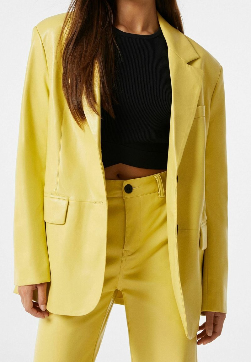 Blazer en cuir jaune avec une coupe décontractée, poche unique, associé à un pantalon jaune assorti et un crop top noir à côtes. Texture lisse.