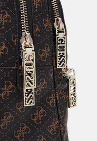 Mustat keinonahkareput, joissa on ruskea monogrammikuosi. Kultaisilla vetoketjuilla, joissa on "GUESS"-logo.